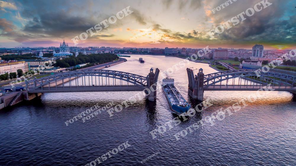 Фотография панорамного вида Санкт-Петербург, Россия, сделанная на закате. На фото запечатлён разведённый Большеохтинский мост, также известный как мост Петра Великого. Фотоснимок передаёт момент навигации на реке Нева, когда пролёты моста подняты для прохода судов. Тёплые оттенки закатного неба подчёркивают силуэт моста и создают выразительную атмосферу вечернего города. Это изображение отражает традицию разводки мостов и панорамный облик Санкт-Петербурга как одного из символов городов России.